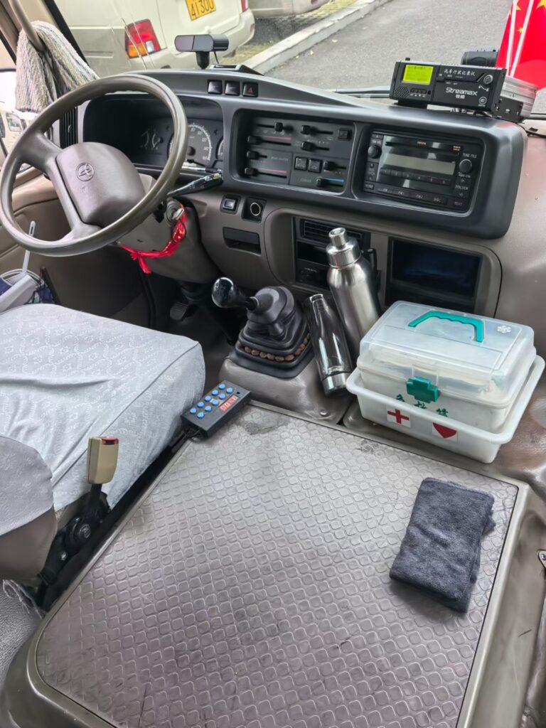 Detailed view of a used Left-Hand Drive (LHD) Toyota Coaster dashboard and driver's cockpit. The image features the original Toyota steering wheel, manual transmission gear shifter, climate controls, and a well-preserved interior. A first-aid kit and professional equipment are visible on the clean engine cover, showing the vehicle is ready for export.