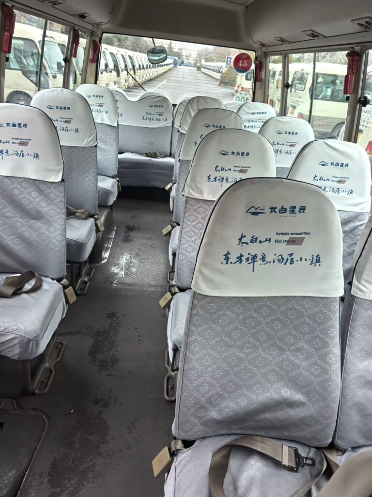 Interior passenger cabin of a used 30-seater Toyota Coaster minibus for export. The image shows multiple rows of well-maintained fabric seats with safety belts and white headrest covers. A clean aisle and large windows are visible, with a fleet of similar buses in the background, showing large inventory at our China yard.