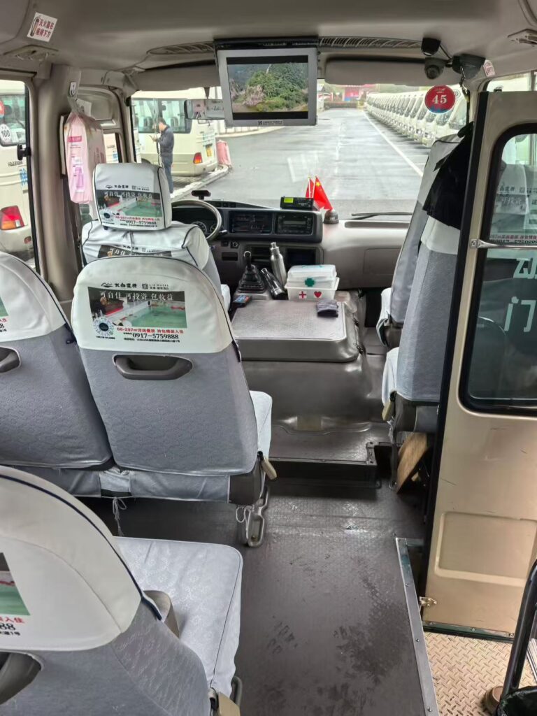 An immaculate, well-preserved interior passenger cabin view of a used Left-Hand Drive (LHD) Toyota Coaster minibus. Tidy grey fabric seats with white headrest covers (featuring some text and graphics), manual transmission controls, and a neat dashboard are visible. Through the windshield, a neat row of similar buses is visible, indicating a large, organized vehicle parking area. The engine cover area is clean, holding a first-aid kit and other items.An immaculate, well-preserved interior passenger cabin view of a used Left-Hand Drive (LHD) Toyota Coaster minibus. Tidy grey fabric seats with white headrest covers (featuring some text and graphics), manual transmission controls, and a neat dashboard are visible. Through the windshield, a neat row of similar buses is visible, indicating a large, organized vehicle parking area. The engine cover area is clean, holding a first-aid kit and other items.